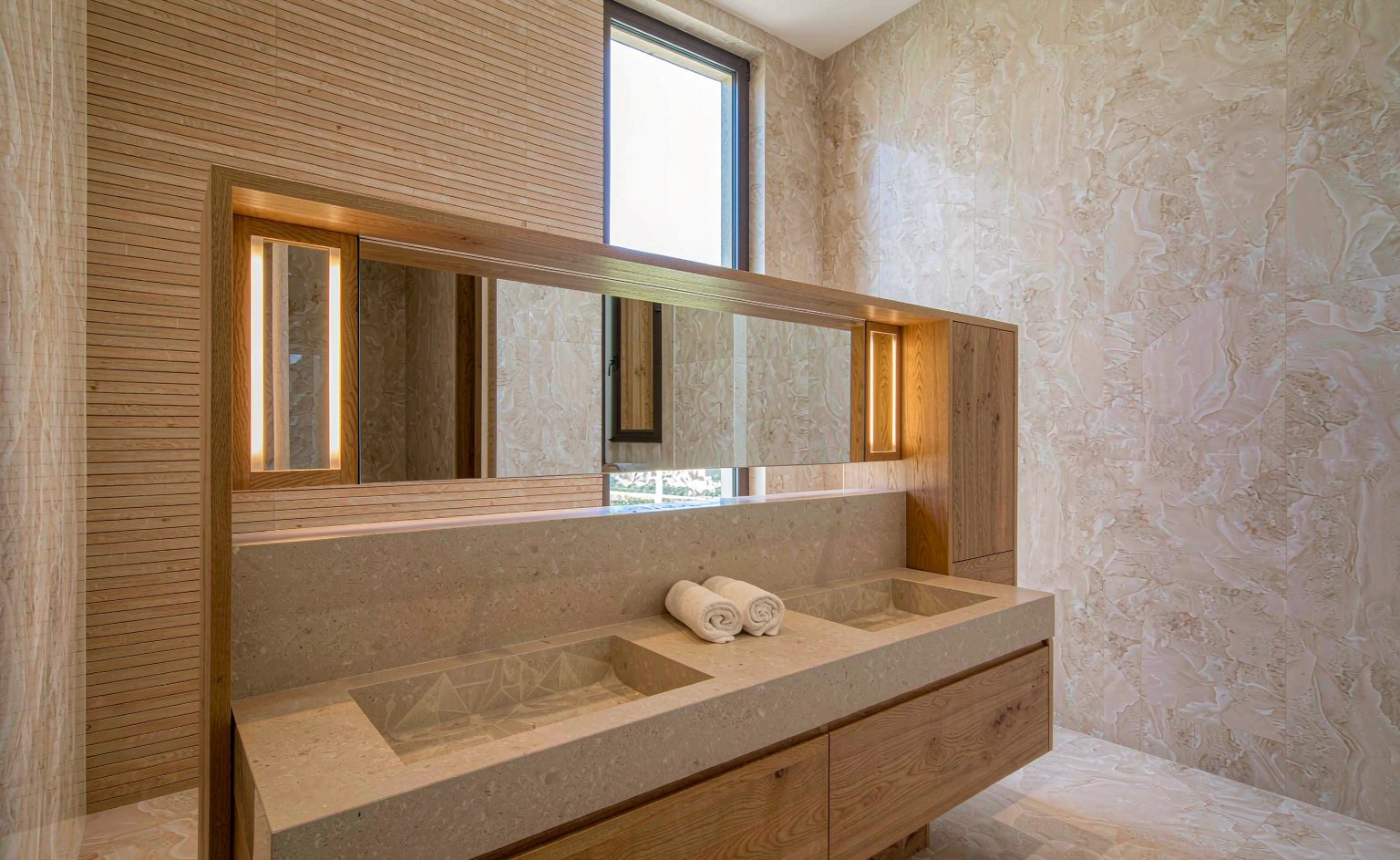 Mueble de baño suspendido de roble con doble lavabo y encimera de piedra, espejo con tiras de luz verticales y pared de listones de madera
