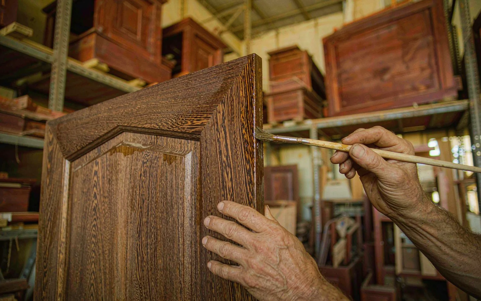 Manos de un artesano aplicando barniz mate con brocha al canto de un panel de madera maciza en un taller de mobiliario